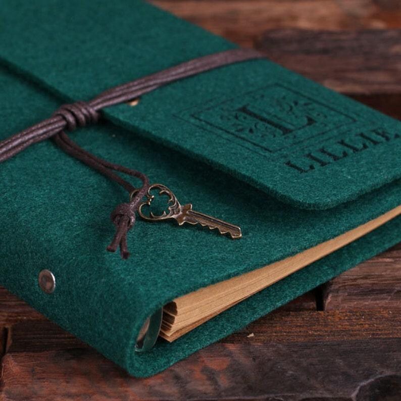 May include: A dark green felt journal with a brown leather strap and a key charm. The journal has a personalized monogram with the letter "I" and the name "Lillie" embossed on the front cover.