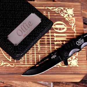 Pu&ograve; includere: Un coltello pieghevole nero con il nome "Ollie" e "Proverbi 18:10" stampati sulla lama. Il coltello &egrave; su una scatola di legno con il nome "Ollie" e "Giovanni 15:12-13" su una placca di metallo e un portafoglio in pelle nera.