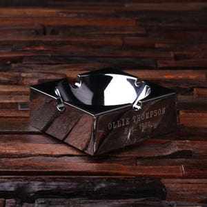 May include: A silver ashtray with a round, recessed center and a square base. The ashtray is engraved with the text "Ollie Thompson Est. 1900."