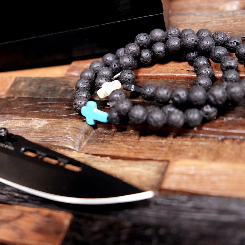 May include: A black lava rock beaded bracelet with a turquoise cross charm and a white bead. The bracelet is lying on a wooden surface.
