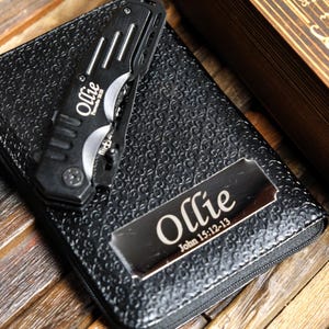Pu&ograve; includere: Un coltello tascabile nero con il nome "Ollie" e il testo "Proverbs 16:30" stampati sul lato, appoggiato su un portafoglio in pelle nero. Una placca metallica sul portafoglio &egrave; incisa con "Ollie John 15:12-13".