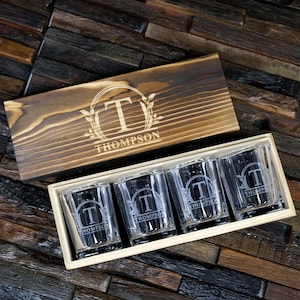 May include: Set of four personalized shot glasses in a wooden box. Each glass features a monogrammed design with a letter "T" inside a circle with a laurel wreath and the name "Thompson" below.