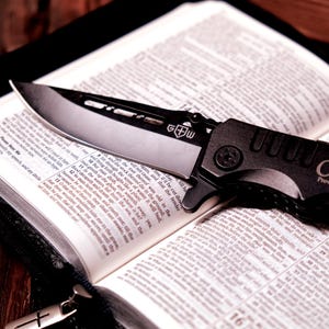 Pu&ograve; includere: Un coltello pieghevole nero e argento con una lama parzialmente seghettata &egrave; appoggiato su un libro aperto. Il coltello ha un'impugnatura strutturata e le lettere "G+W" sulla lama. Le pagine del libro sono piene di testo.