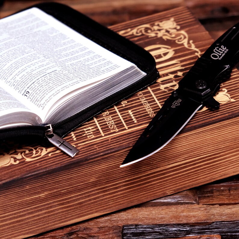 Pu&ograve; includere: Un coltello pieghevole nero con la scritta "Ollie" sulla lama &egrave; appoggiato su una superficie di legno accanto a una Bibbia aperta in una custodia nera. La Bibbia &egrave; aperta su una pagina con testo. La superficie in legno presenta intagli decorativi.