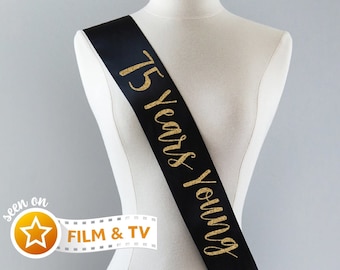 May include: Black satin sash with gold glitter lettering that reads "75 Years Young".
