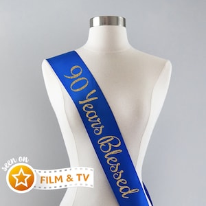 May include: Blue satin sash with gold glitter lettering that reads "90 Years Blessed".