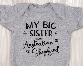 Könnte beinhalten: Grauer Baby-Strampler mit schwarzem Text, der "Meine große Schwester ist ein Australian Shepherd" lautet, mit einer Pfotenabdruckgrafik.