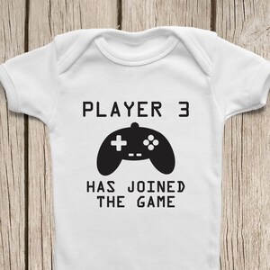 May include: White baby onesie with black text that reads "Player 3 Has Joined The Game" and a black video game controller graphic.