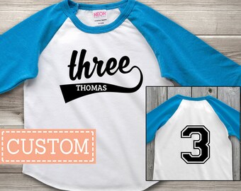 May include: A white and blue raglan style t-shirt with the word "three" in black script and the name "Thomas" below. The shirt has a custom design and a number 3 on the back.