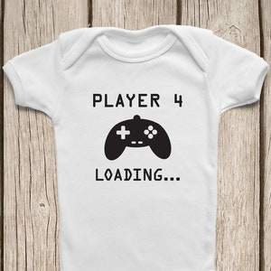 May include: White baby onesie with the text "PLAYER 4" above a black video game controller graphic and the word "LOADING..." below. The onesie is made of soft material and has snap closures.