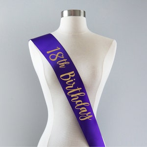 May include: A purple satin sash worn diagonally across a mannequin torso. The sash features the text 18th Birthday printed in a gold glitter script font that runs vertically from the shoulder down to the waist area of the display form.