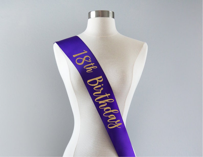 May include: A purple satin sash worn diagonally across a mannequin torso. The sash features the text 18th Birthday printed in a gold glitter script font that runs vertically from the shoulder down to the waist area of the display form.