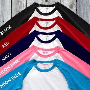 May include: A collection of baseball-style shirts with white bodies and colored sleeves. Colors include black, red, navy, neon pink, and neon blue. Each sleeve has the color name printed in white.