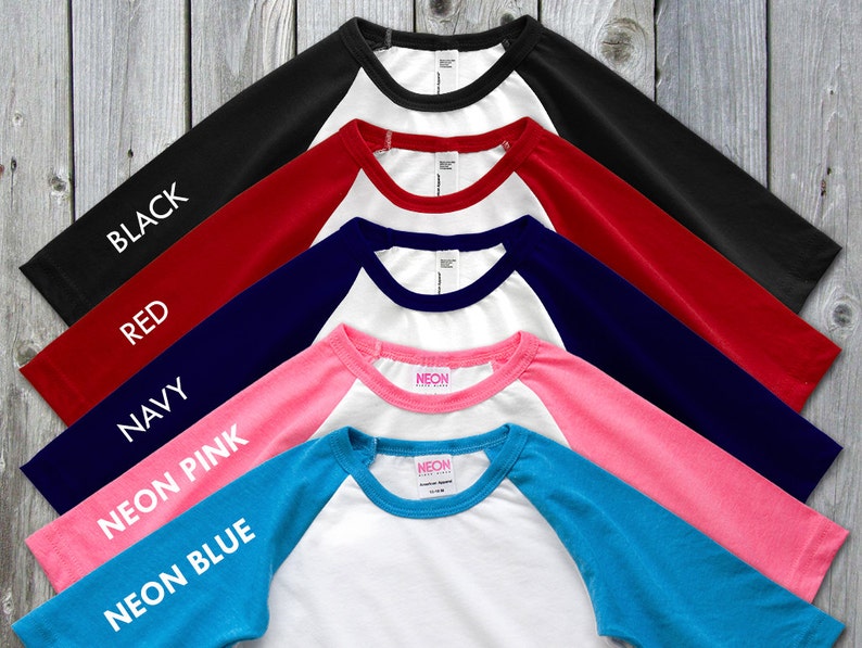 May include: A collection of baseball-style shirts with white bodies and colored sleeves. Colors include black, red, navy, neon pink, and neon blue. Each sleeve has the color name printed in white.