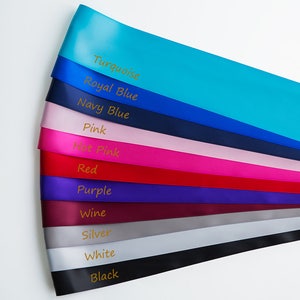 May include: A collection of ten satin ribbon rolls in various colors, including turquoise, royal blue, navy blue, pink, hot pink, red, purple, wine, silver, white, and black. Each roll has the color name printed on it in gold lettering.