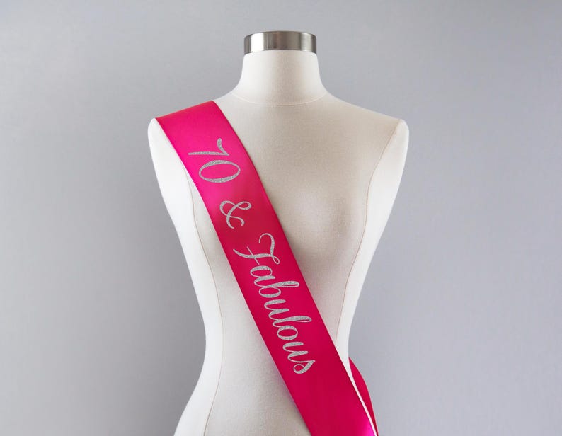 Pu&ograve; includere: Una fascia in raso rosa acceso drappeggiata su un busto di manichino bianco. La fascia presenta la scritta 70 & Fabulous in un carattere corsivo glitterato color argento che corre in diagonale dalla spalla fino alla vita del manichino.