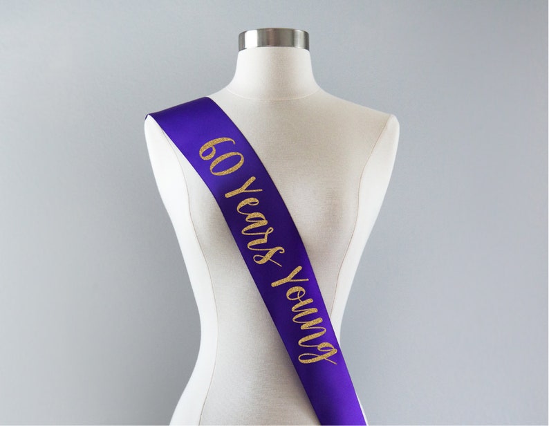 Pode incluir: Faixa de cetim roxo com letras douradas de glitter que dizem "60 Years Young".