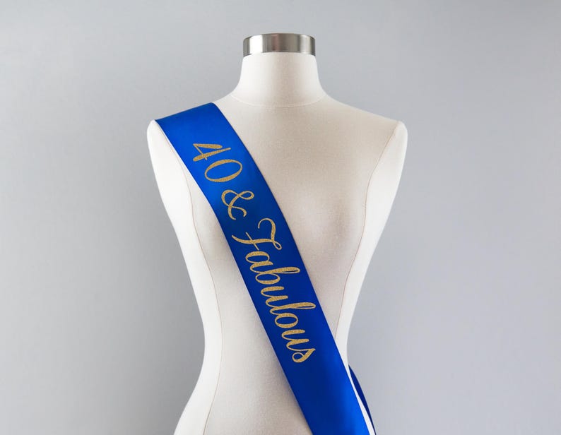 May include: A royal blue satin sash draped across a white mannequin torso. The sash features the text 40 & Fabulous printed in a gold glitter script font, running diagonally from the shoulder down to the waist area of the display form.