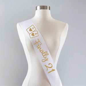 May include: A white satin sash featuring gold glitter text that reads Finally 21, accompanied by a graphic of two playing cards showing a Queen and an Ace of hearts. The sash is displayed on a neutral-colored mannequin torso against a plain background.
