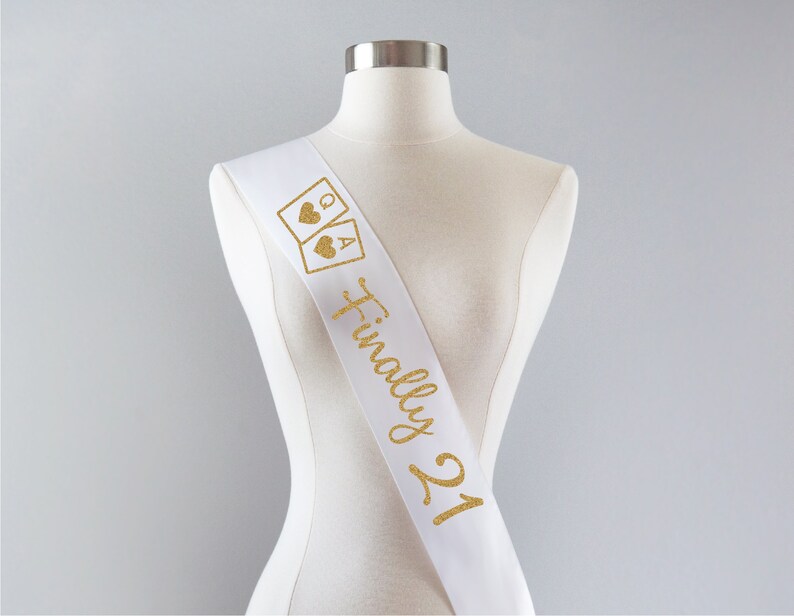 May include: A white satin sash featuring gold glitter text that reads Finally 21, accompanied by a graphic of two playing cards showing a Queen and an Ace of hearts. The sash is displayed on a neutral-colored mannequin torso against a plain background.