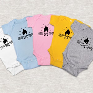 Op de afbeelding: Vijf baby-rompers in verschillende kleuren met een zwart-witte afbeelding van een kampvuur en de tekst "Happy Camper". De rompers zijn wit, lichtblauw, roze, geel en grijs.