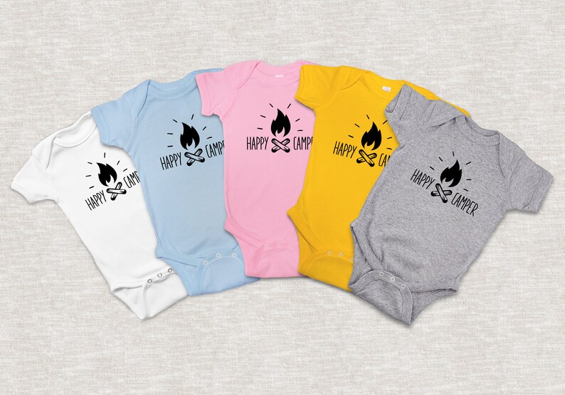 Op de afbeelding: Vijf baby-rompers in verschillende kleuren met een zwart-witte afbeelding van een kampvuur en de tekst "Happy Camper". De rompers zijn wit, lichtblauw, roze, geel en grijs.