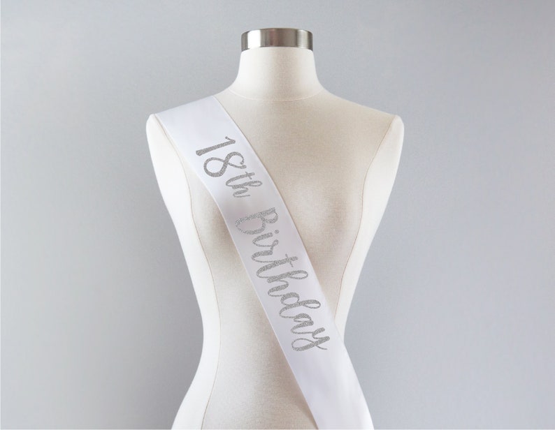 May include: A white satin sash featuring the text 18th Birthday written in a silver glitter script font. The sash is draped diagonally across a mannequin torso, displaying the celebratory lettering clearly against the smooth white fabric surface.
