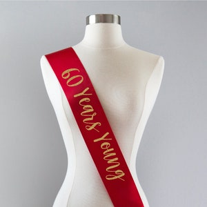 Pode incluir: Faixa de cetim vermelho com letras douradas brilhantes que dizem "60 Years Young".