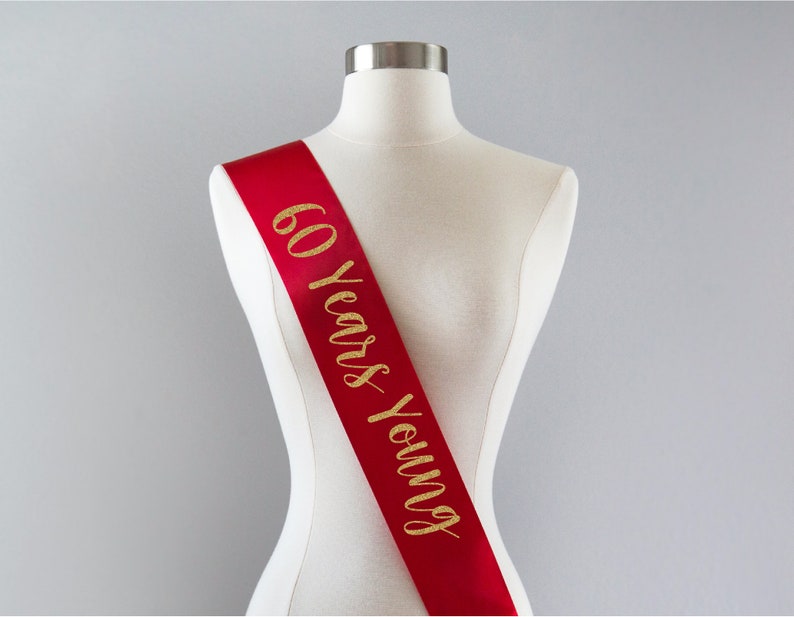 Pode incluir: Faixa de cetim vermelho com letras douradas brilhantes que dizem "60 Years Young".