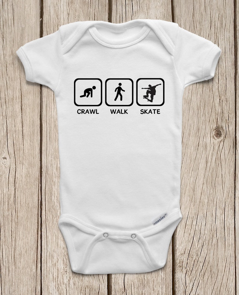 May include: White baby onesie with black text and illustrations. The text reads "Crawl, Walk, Skate" and there are three icons depicting a person crawling, walking, and skateboarding.