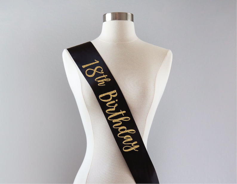 May include: A black satin sash featuring the text 18th Birthday written in a gold glitter script font. The sash is draped diagonally across a neutral-colored mannequin torso, displaying the celebratory lettering clearly against the dark fabric.