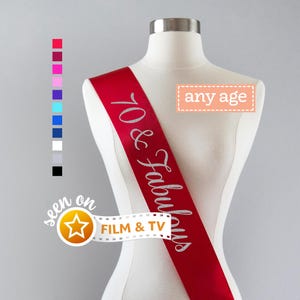 Pu&ograve; includere: Una fascia in raso rosso indossata su un busto di manichino, con la scritta in glitter argento 70 & Fabulous. Un riquadro rettangolare contiene il testo any age, e una grafica a pellicola include il testo seen on FILM & TV con una stella.