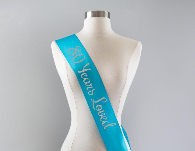 May include: A bright turquoise satin sash draped across a white mannequin torso. The sash features the text 80 Years Loved written in a cursive, glittery silver font that runs vertically along the length of the fabric from shoulder to hip.