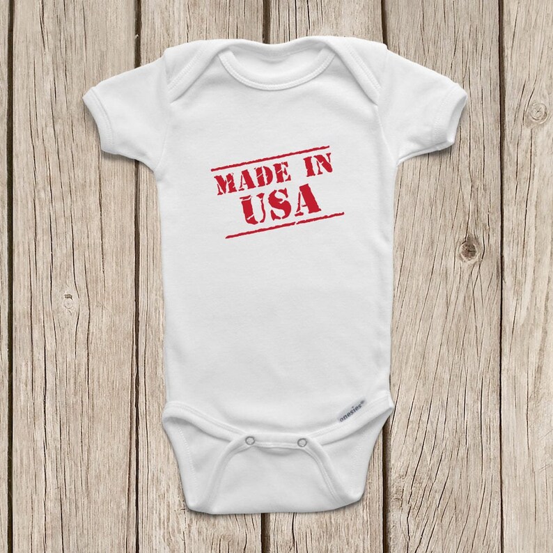 Puede incluir: Body de beb&eacute; blanco con el texto rojo "MADE IN USA" estampado en el pecho. El body tiene mangas cortas y corchetes en la parte inferior. El fondo es una superficie de madera marr&oacute;n claro.