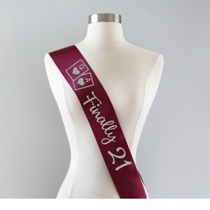 May include: A deep burgundy satin sash featuring silver glitter text that reads Finally 21, accompanied by a graphic of two playing cards, a queen and an ace, each displaying a heart symbol, displayed on a neutral-colored mannequin torso.