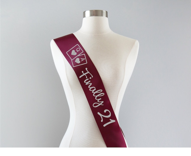 May include: A deep burgundy satin sash featuring silver glitter text that reads Finally 21, accompanied by a graphic of two playing cards, a queen and an ace, each displaying a heart symbol, displayed on a neutral-colored mannequin torso.