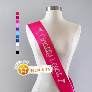 May include: A bright pink satin sash worn on a mannequin, featuring the silver glitter text Finally Legal with martini glass icons at each end. A vertical column of twelve small colored squares is visible on the left, along with a film strip graphic.