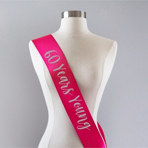 Pode incluir: Faixa de cetim rosa com letras de glitter prata que diz "60 Years Young".