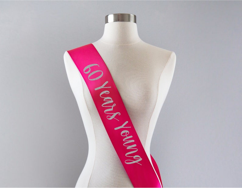 Pode incluir: Faixa de cetim rosa com letras de glitter prata que diz "60 Years Young".