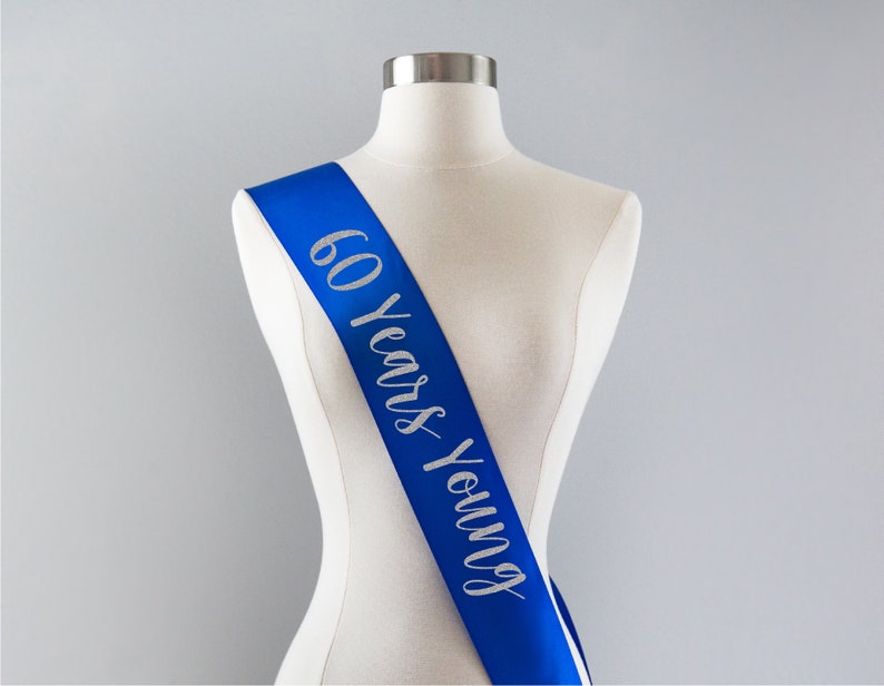 Pode incluir: Uma faixa de cetim azul com letras de glitter prateadas que dizem "60 Years Young".