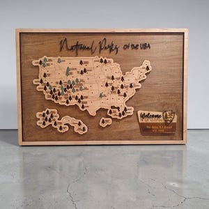 May include: A rectangular wooden wall map of the USA featuring laser-cut National Parks of the USA text at the top. The map includes small tree icons marking locations and a plaque in the bottom right corner that reads Welcome to Our Adventure.