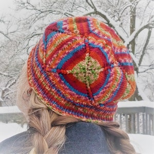 May include: A colorful, hand-knitted hat with a geometric design. The hat features a vibrant mix of red, orange, blue, and green yarns. The hat is worn by a person with blonde hair in a braid, with a snowy background.