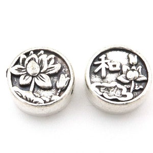 May include: Two silver-toned round beads with raised designs. One bead features a lotus flower and the other bead features a lotus flower and the Chinese character for "peace".