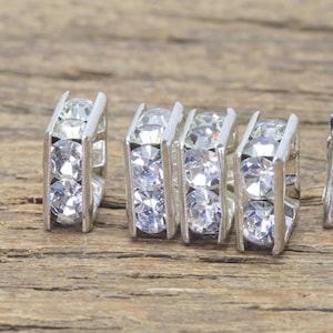 May include: Five silver-toned square-shaped earring settings with clear rhinestones. The settings are arranged in a row on a wooden surface.