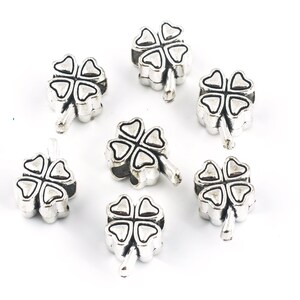 May include: Silver-colored clover charms with four leaves, arranged on a white background. Each charm has a detailed design, with visible lines defining the leaves and stem. These charms are suitable for jewelry making or craft projects.