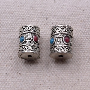 May include: Two silver cylindrical beads with intricate designs and red and blue gemstones. The beads feature the Om symbol.