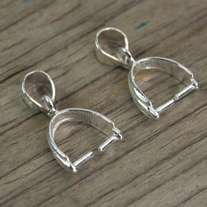 May include: Two silver-toned pendant bails with a loop at the top and a curved opening at the bottom. The bails are designed to hold a pendant or charm.