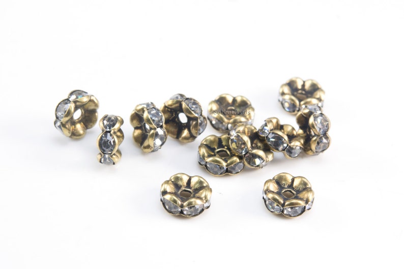 May include: A group of antique gold-toned metal beads with clear rhinestone accents. The beads are round and have a flower-like design.