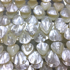May include: A close-up of a pile of white, iridescent, shell-shaped beads. The beads are smooth and have a pearlescent finish.