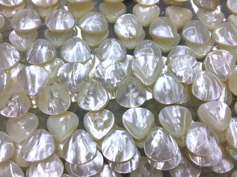 May include: A close-up of a pile of white, iridescent, shell-shaped beads. The beads are smooth and have a pearlescent finish.